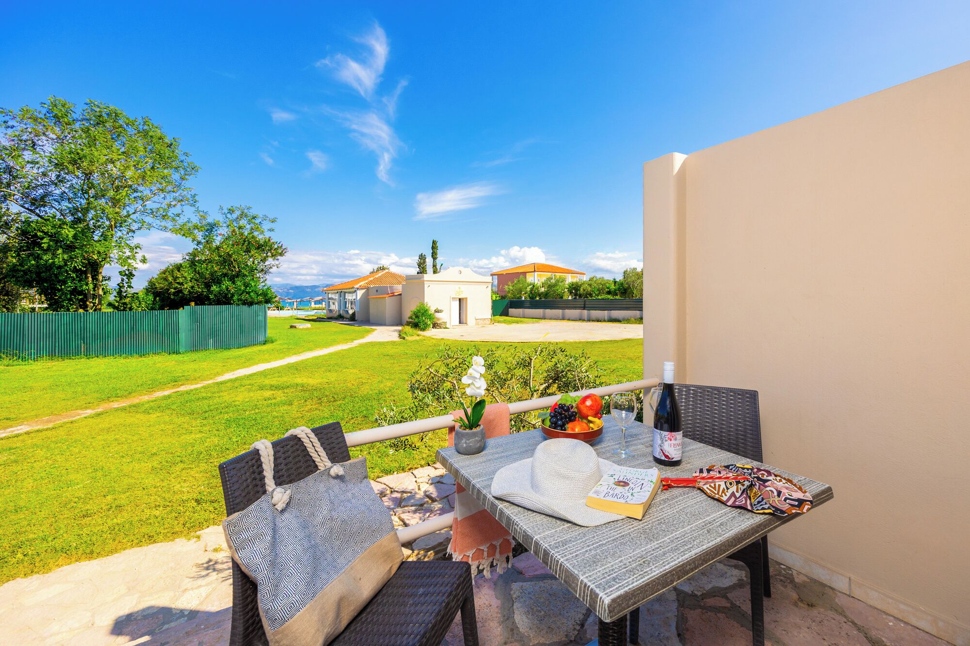 A patio with a table set for dining, a view of a pool and buildings, and a green fence in the background.