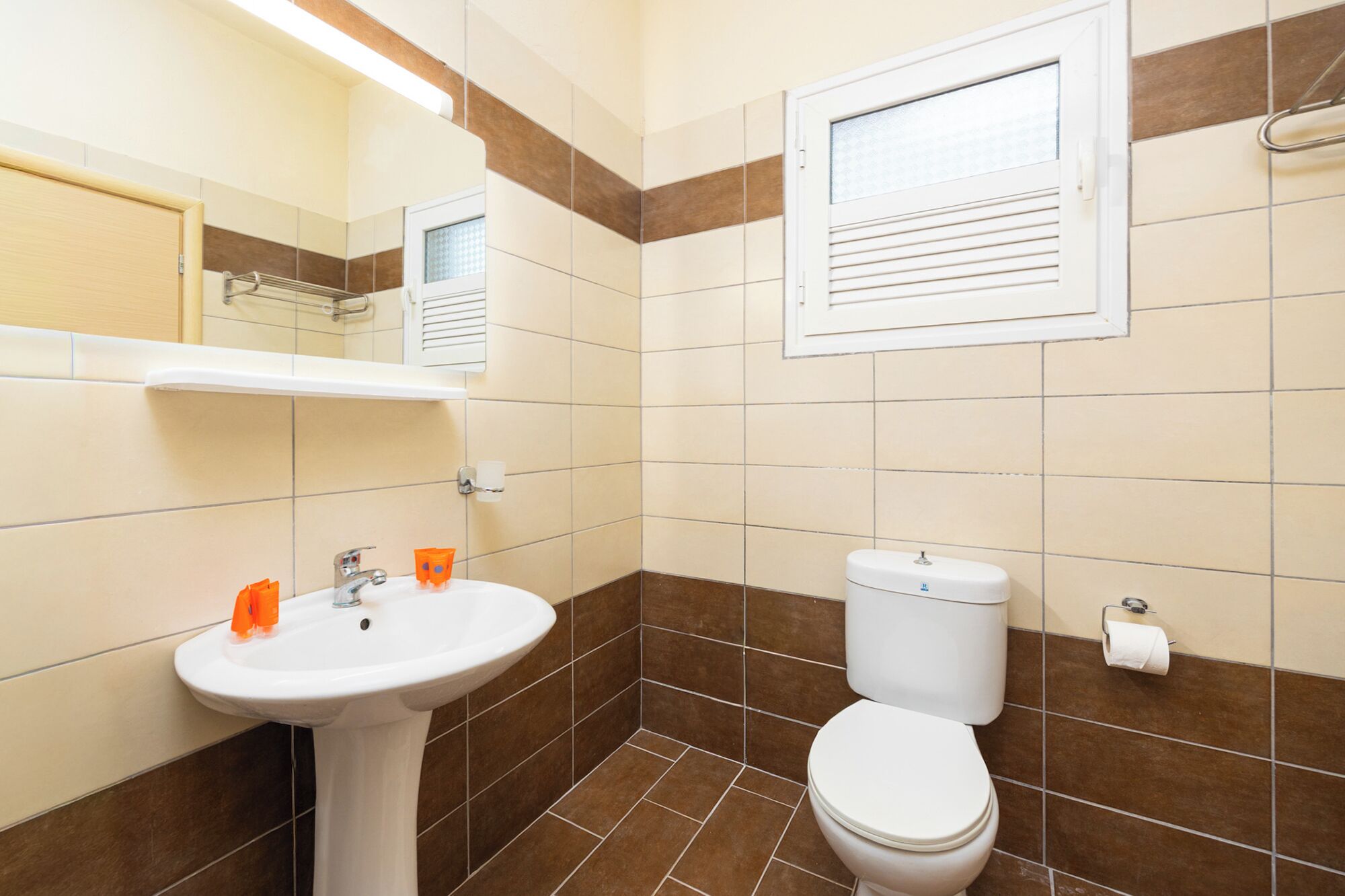 Een badkamer met een wastafel op voetstuk, een toilet en een raam met zonwering.