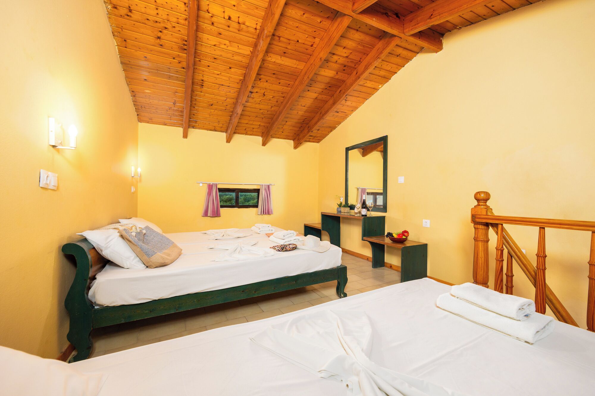 A room with a wooden ceiling, a bed with a green frame, a bedside table with a mirror, and a wooden staircase.