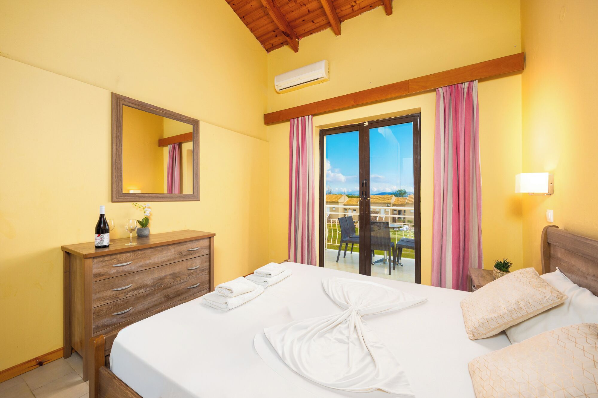 A hotel room with a bed, a wooden dresser, a mirror, a window with curtains, and a balcony with chairs.