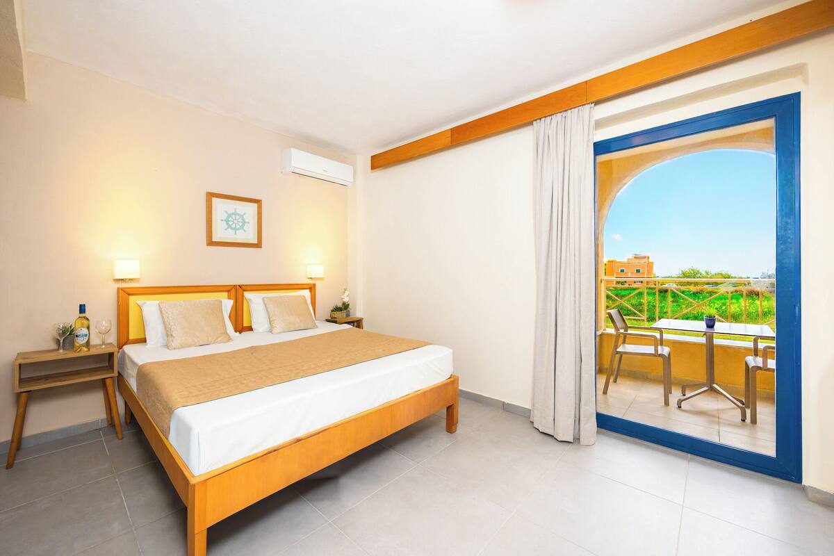A hotel room with a bed, bedside tables, a framed picture, an air conditioning unit, and a balcony with a table and chairs.
