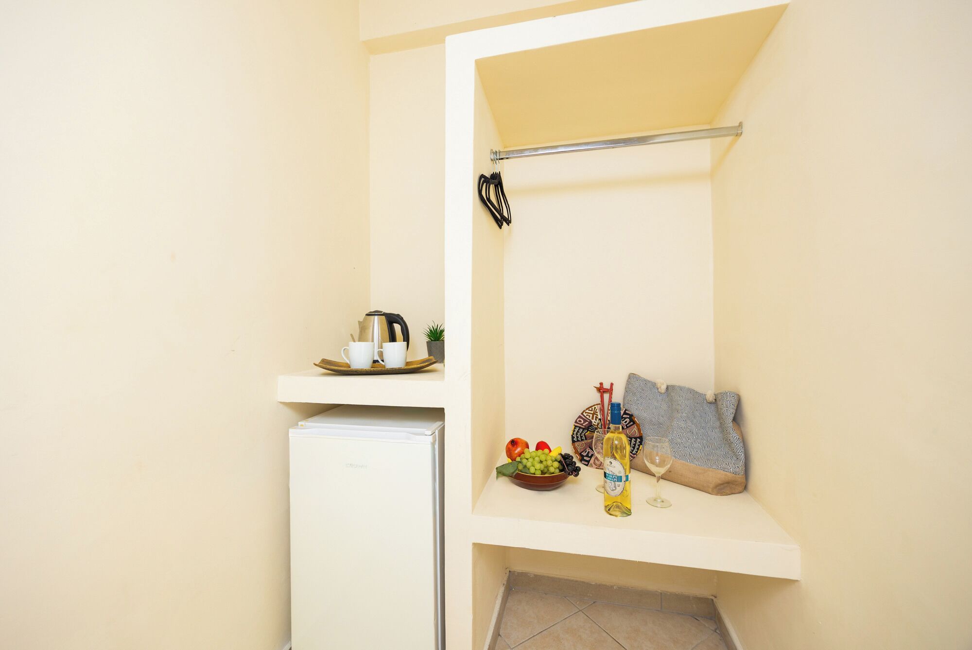 A compact kitchenette with a refrigerator, a small shelf with a kettle, cups, and a tray, and a bench with a bottle and a glass.