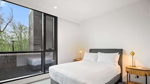 A modern bedroom with a large window, a bed with white linens, two bedside tables with lamps, and a view of trees outside. - Brand-New 2-Bed with Rooftop Access (Northcote)