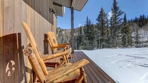 A wooden deck with two Adirondack chairs, a wooden building with a sloped roof, and a snowy landscape with trees in the background. - Stunning Views from a Unique Mountain Cabin! (Jefferson)