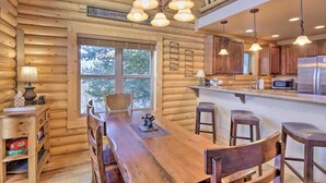 A log cabin interior with a wooden dining table, bar stools, a kitchen area with a refrigerator, and a staircase.