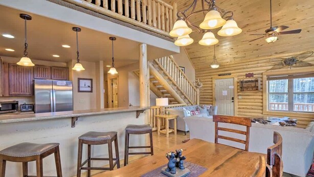 A spacious log cabin interior with a wooden dining table, bar stools, a kitchen with stainless steel appliances, and a staircase leading to a loft area.