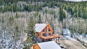 A log cabin with a large deck and a separate building, surrounded by a forested area with snow on the ground.