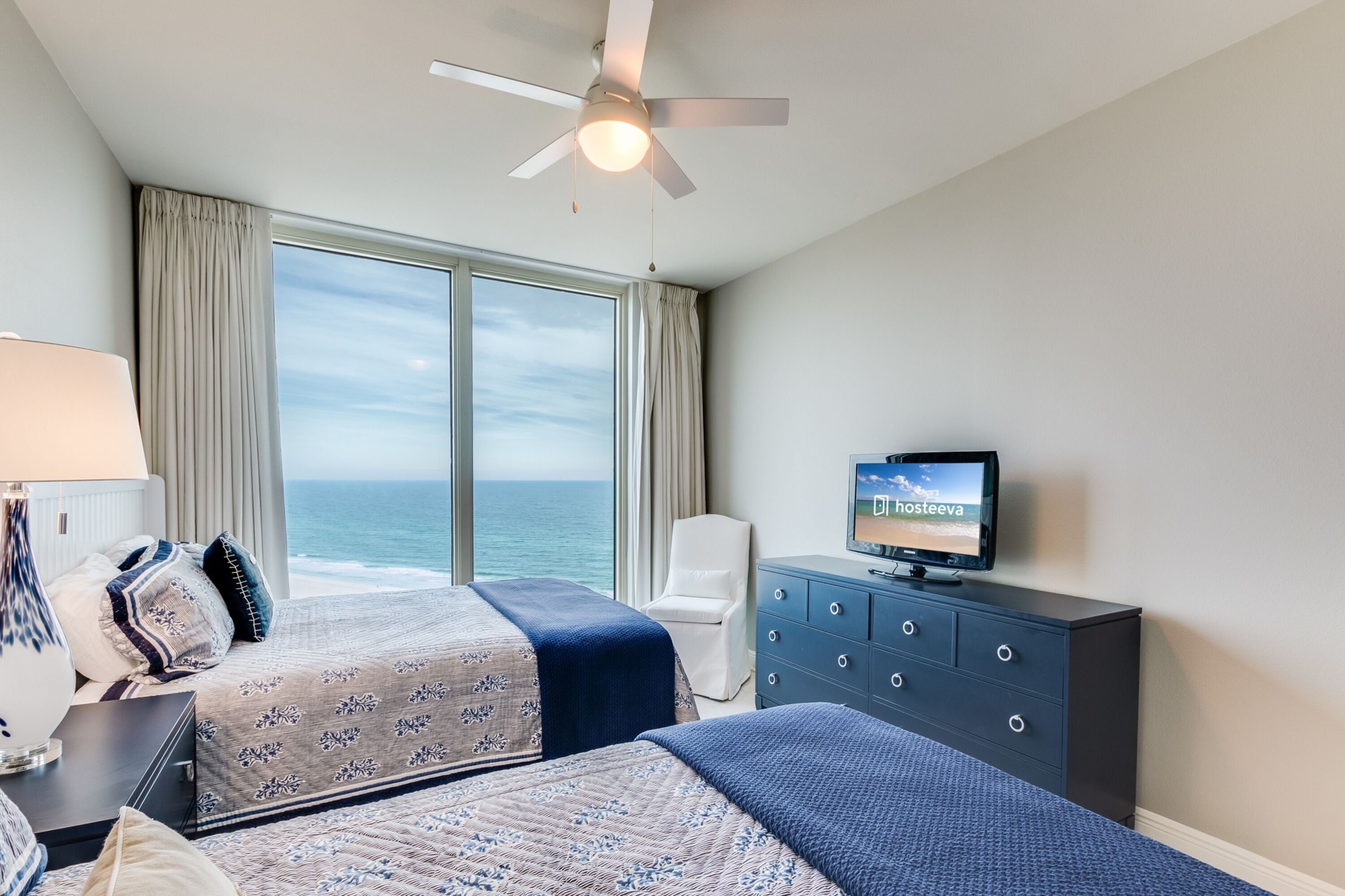Een hotelkamer met twee bedden, een televisie, een bureau met een lamp, een stoel en uitzicht op de oceaan.
