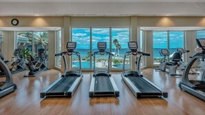 Academia com esteiras, bicicletas ergométricas e elípticos, amplas janelas com vista para o mar.