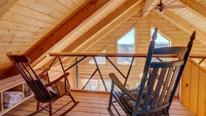 Un interior de madera con techo inclinado, dos mecedoras, una mesa y un ventanal con vistas al paisaje.