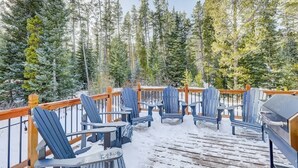 Un terrazzo in legno con poltrone Adirondack e un barbecue, circondato da alti alberi sempreverdi.