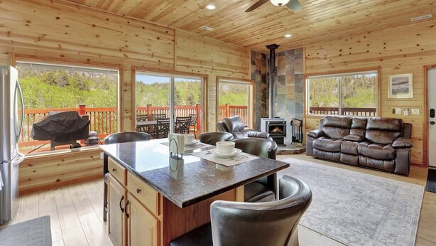 Interior de una cabaña de madera con cocina, chimenea, sofá de cuero y amplias ventanas que ofrecen vistas al exterior.
