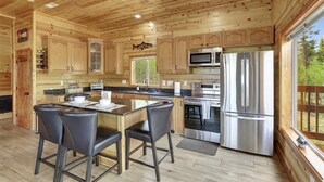 Una cocina amplia con armarios de madera, una isla central con taburetes de bar, electrodomésticos de acero inoxidable y un amplio ventanal con vistas al exterior.
