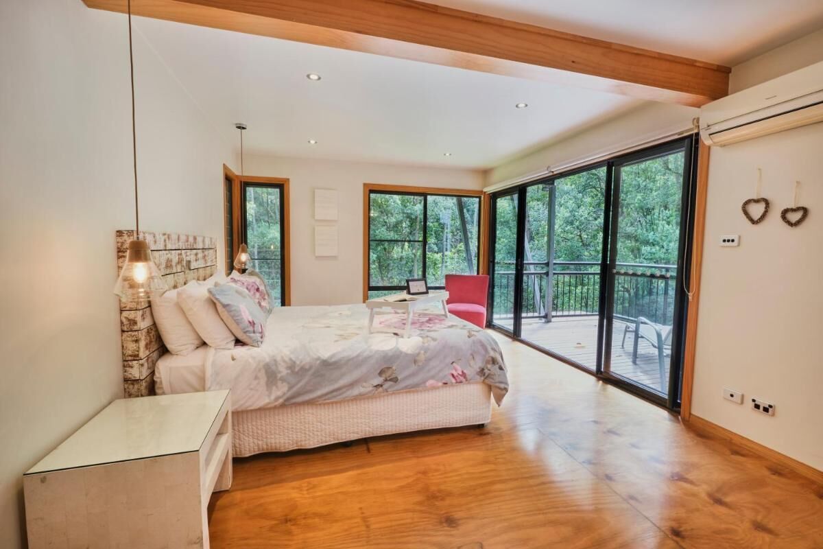 A bedroom with a large bed, a nightstand, a red chair, and a balcony with a view of trees.