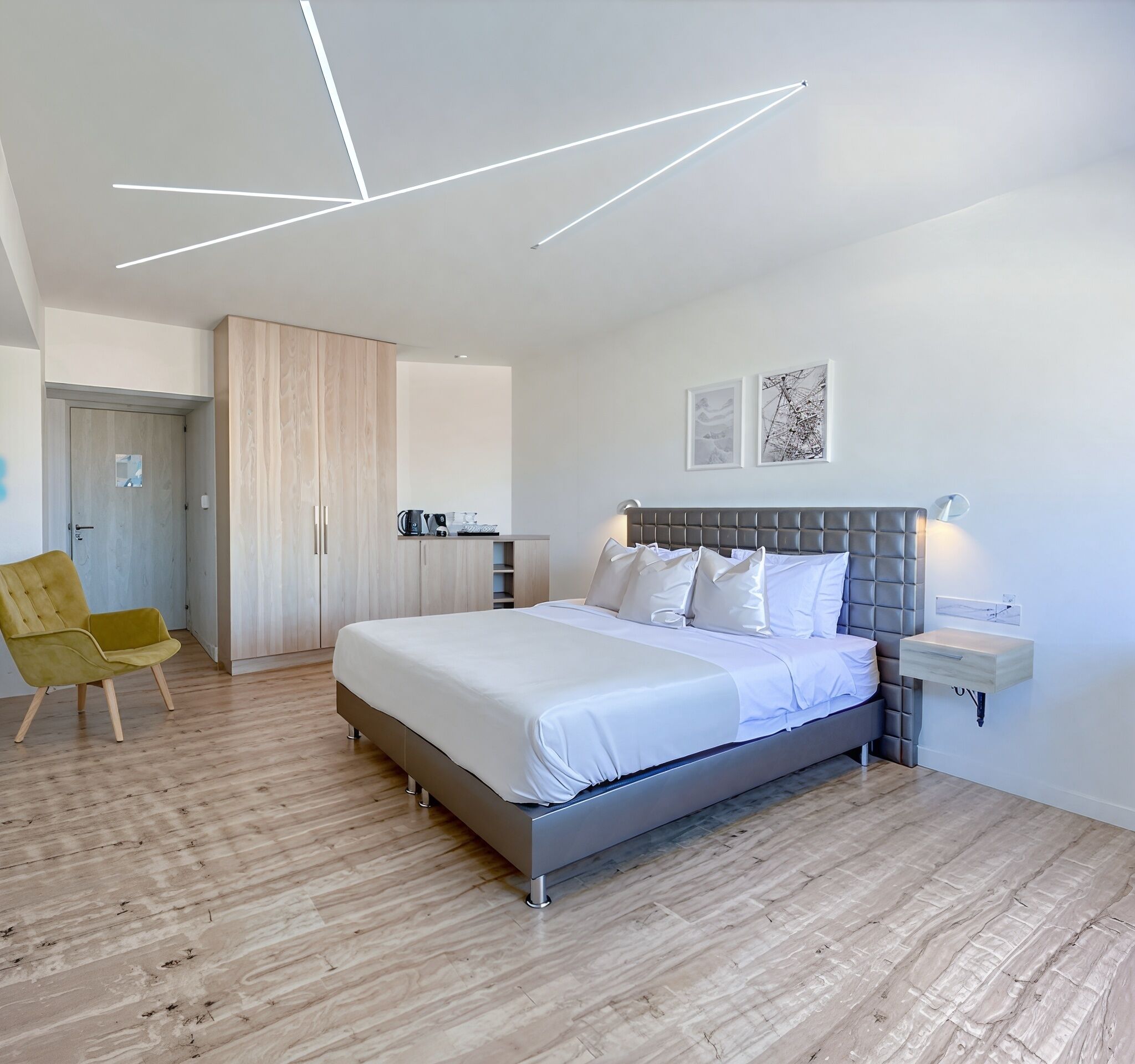 A modern bedroom with a large bed, a chair, a wardrobe, and a small kitchenette.