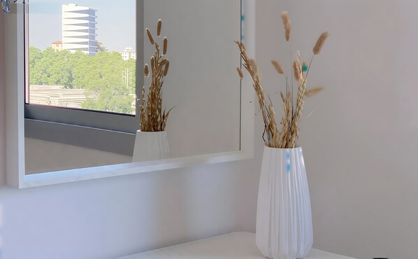 Un jarrón blanco con plantas secas sobre una superficie blanca, junto a una ventana que ofrece vistas a edificios y árboles.