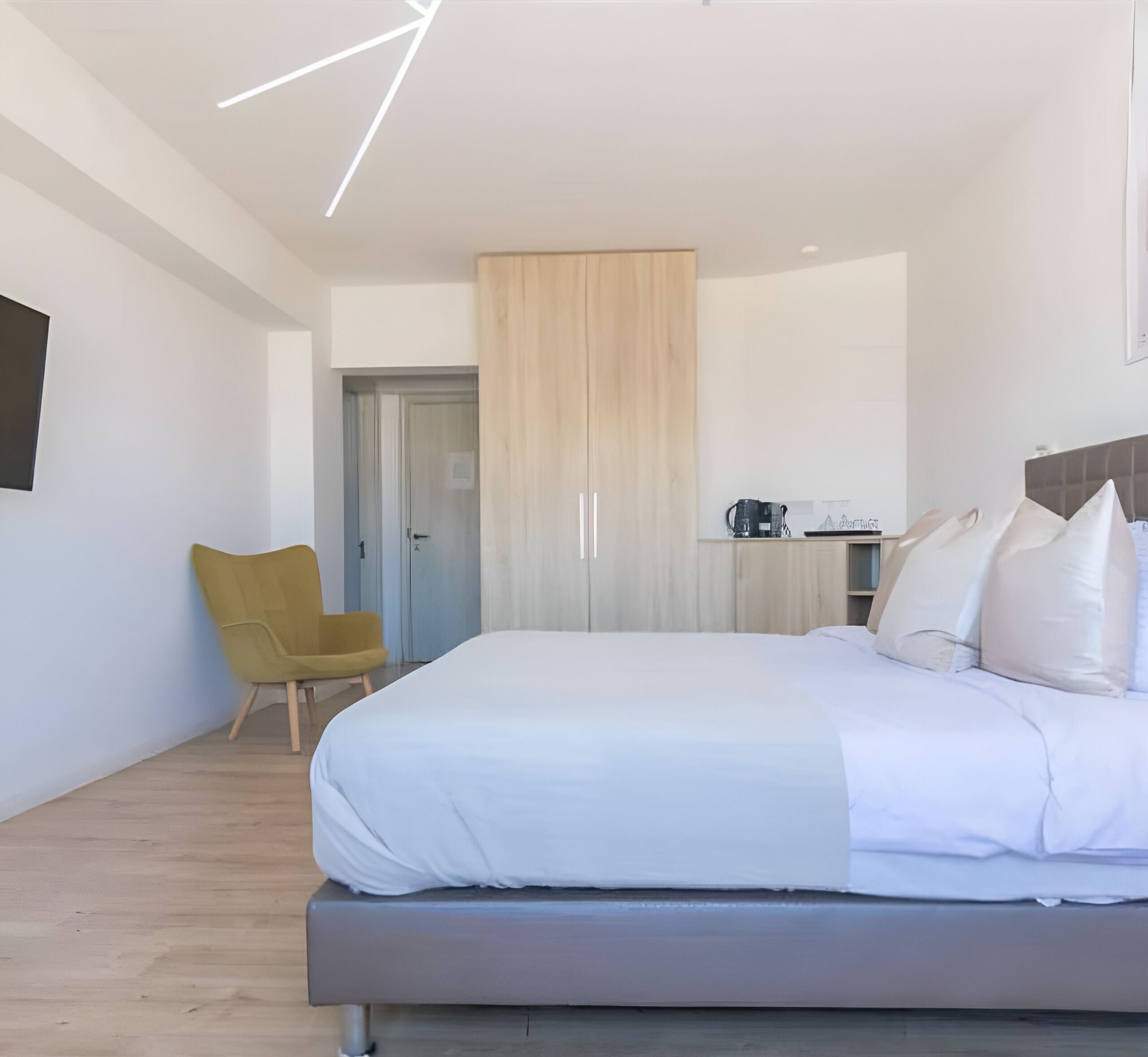 Una habitación de hotel moderna con una cama grande, un armario de madera, un escritorio con cafetera y un sillón amarillo.