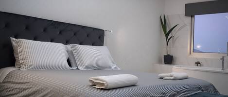 A modern bedroom with a tufted headboard, striped bedding, a neatly folded white towel, and a potted plant by the window.