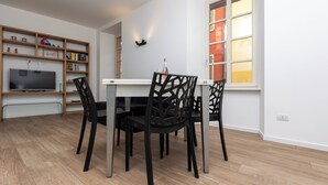 Un comedor con una mesa blanca, sillas negras, un televisor en un estante de madera y una ventana con cortinas naranjas.