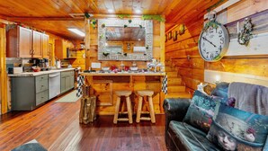 L’intérieur d’un chalet rustique, avec des murs en bois, un plafond en bois, un coin cuisine doté d’un évier et de placards, une table à manger avec des tabourets et un canapé agrémenté de coussins décoratifs.