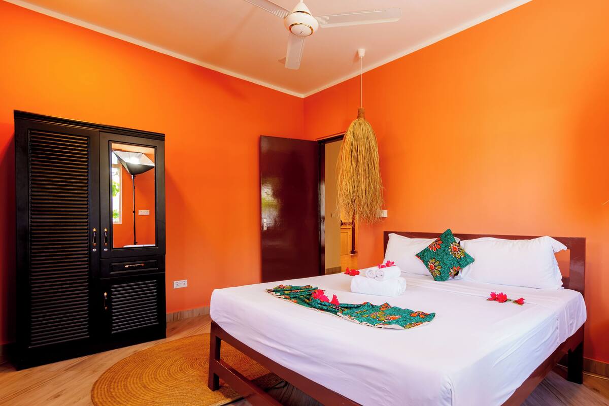 Une pièce avec un mur orange, un lit recouvert de draps blancs, un luminaire suspendu et une porte en bois.