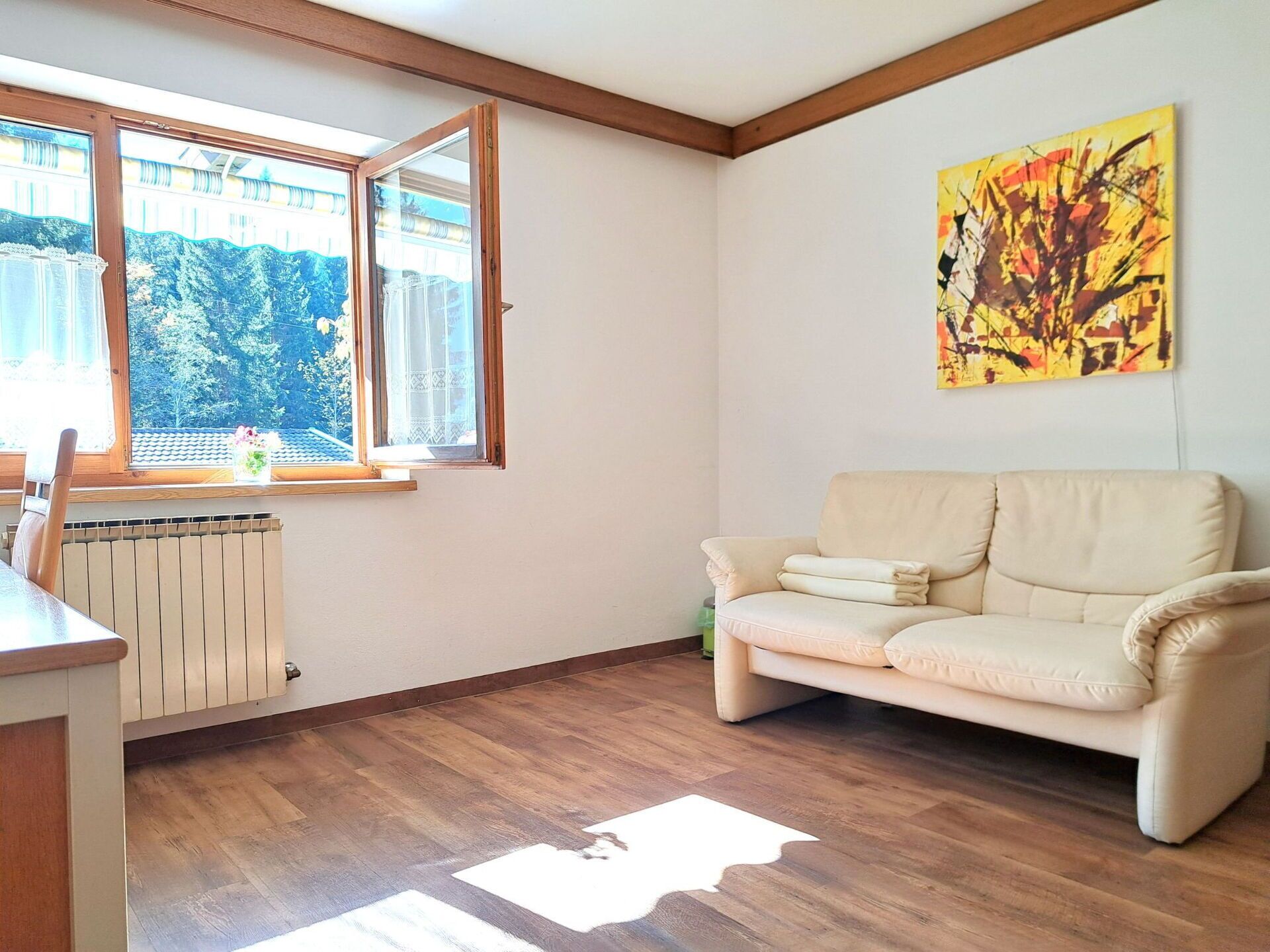 A living room with a beige sofa, a radiator, a window with a view of trees, and a painting on the wall.