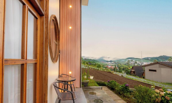 A balcony with a table and chairs, large windows, and a view of a landscape with buildings and hills. - Umacabana 2 (Dieng Kulon)