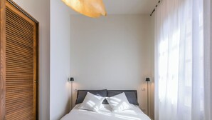 A neatly made bed with a white bedspread, two pillows, and a headboard. There are two bedside lamps and a window with sheer curtains on the right.