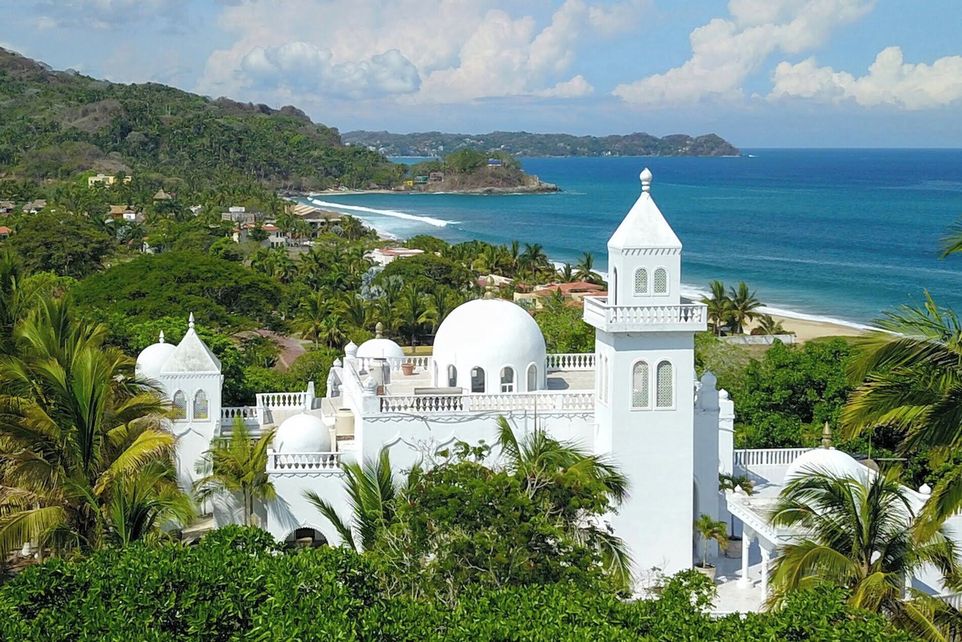 A white building with multiple domes and towers, surrounded by lush greenery and overlooking a beach and ocean.