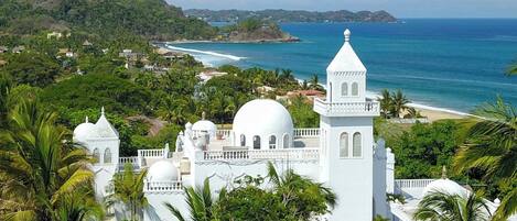 A white building with multiple domes and towers, surrounded by lush greenery and overlooking a beach and ocean.