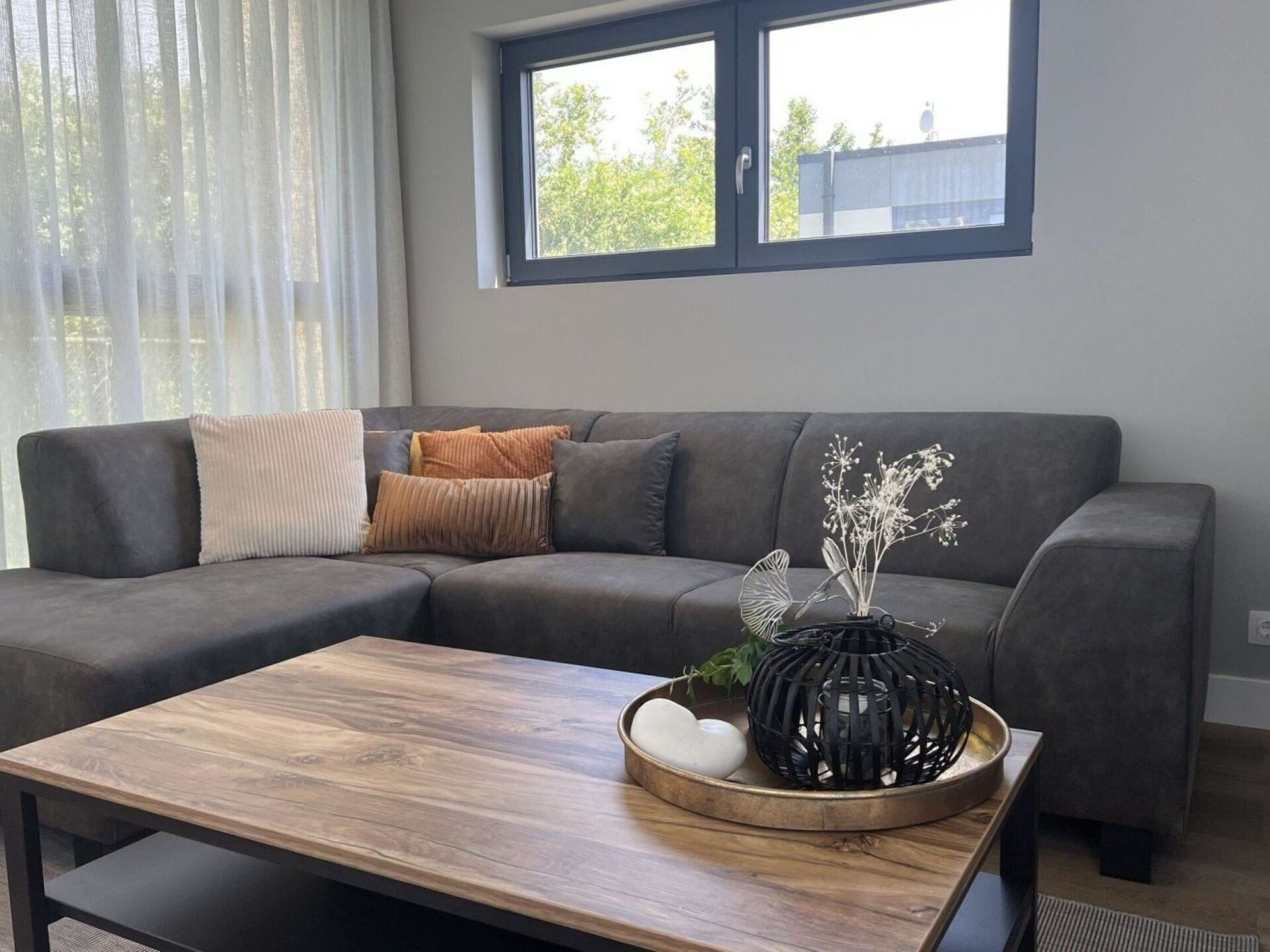 Un salon moderne avec un canapé d’angle gris, une table basse en bois et une fenêtre équipée de stores.