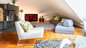 Un loft modern amb una llar de foc, un televisor gran que mostra 'NETFLIX' i una zona de seients acollidora amb una catifes estampada.