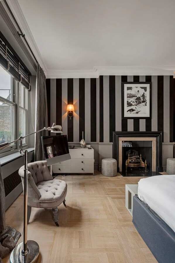 A bedroom with striped wallpaper, a fireplace, a bed, a desk with a lamp, and a chair. - Cadogan Squre-2 Bedroom (London)