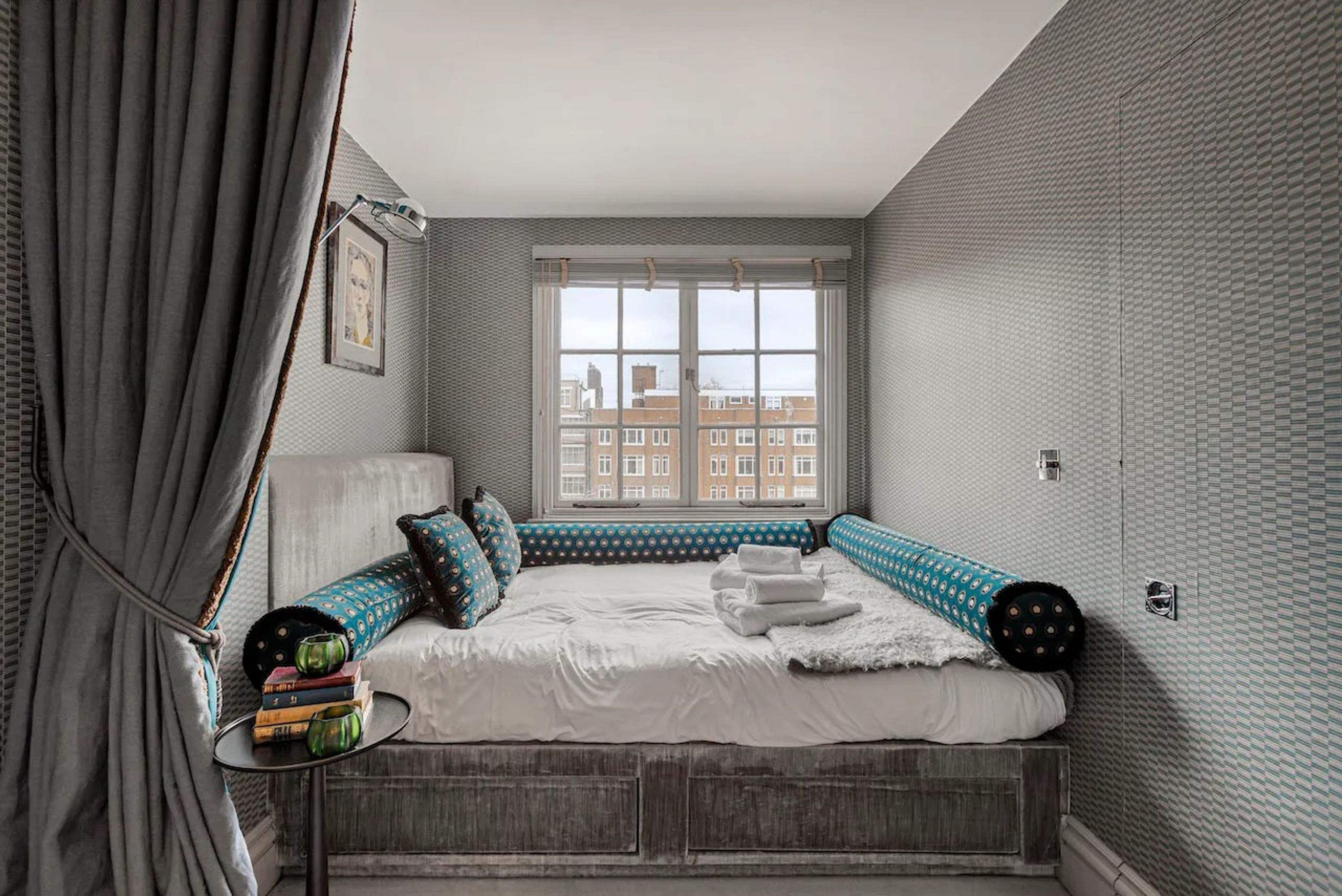 A bedroom with a large window, a bed with blue and white bedding, and a view of buildings outside.