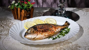 A grilled fish on a plate with lemon slices and parsley, set on a table with a lace tablecloth.