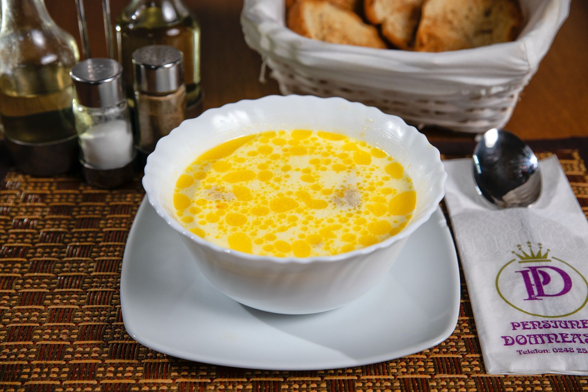Un bol de soupe blanche sur une assiette blanche, accompagné de salière et de poivrière, d’un panier de pain et d’une serviette décorée d’un logo.