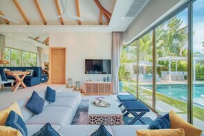 A modern living room with a large TV, comfortable seating, and a view of a swimming pool. - Stylish 3BR Pool Villa, Bang Tao + Airport Shuttle (Choeng Thale)