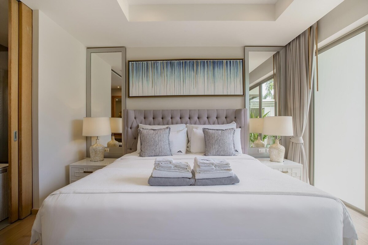 A neatly made bed with a tufted headboard, two bedside tables with lamps, and a large window with curtains.