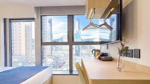 Ein modernes Hotelzimmer mit einem großen Bett, einem Schreibtisch, einem Stuhl, einem an der Wand befestigten Fernseher und Blick auf die Stadt durch große Fenster.