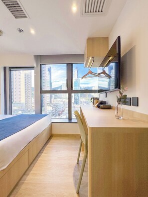 A modern hotel room with a large bed, a desk, a chair, a TV mounted on the wall, and a view of the city through large windows. - Bluejay Residences Kai Tak (Kowloon)