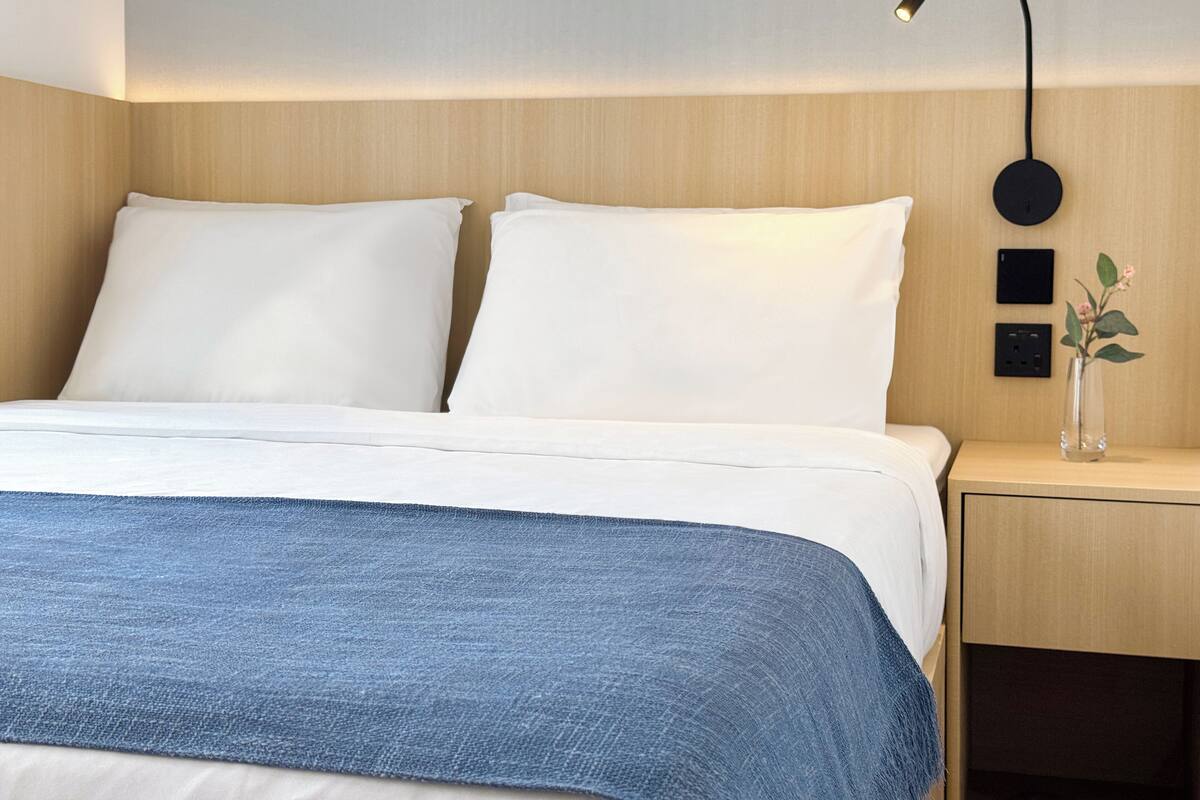 A neatly made bed with white linens and a blue blanket, a bedside table with a vase of flowers, and a wall-mounted lamp.