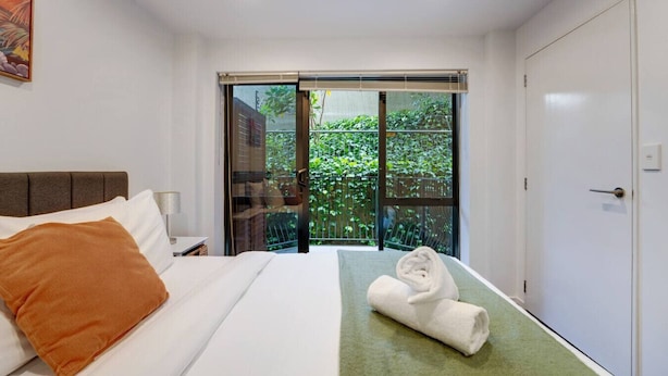 A modern hotel room with a bed, a nightstand, a window with a view of greenery, and a sliding door to a balcony.