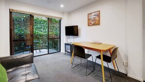 A room with a sliding glass door leading to a balcony, a wooden table with chairs, a flat-screen TV, and a painting on the wall.
