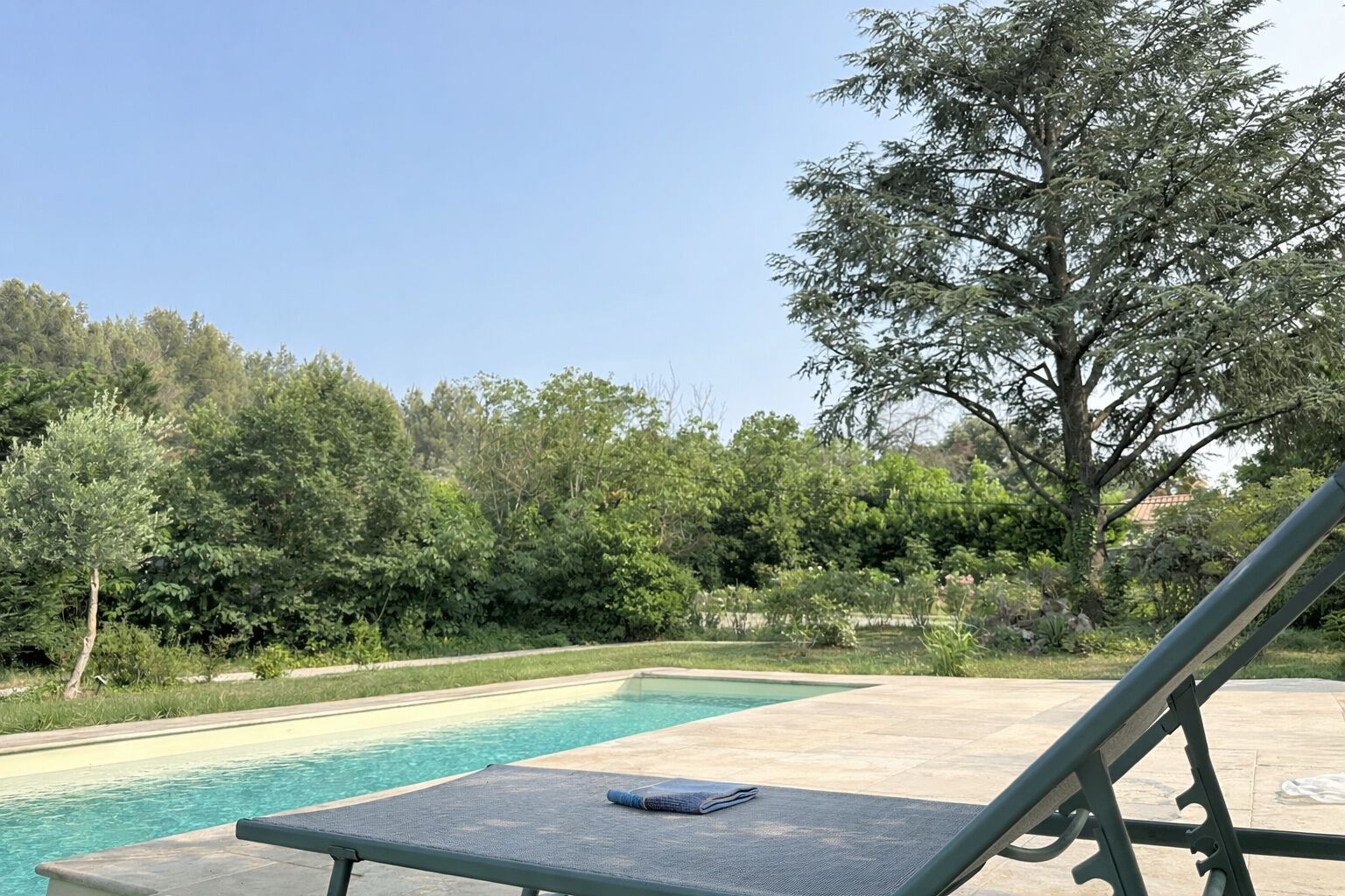 Un espace aménagé au bord de la piscine, avec un fauteuil de relaxation et une serviette, entouré d’une végétation luxuriante et d’arbres.