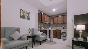 Un espacio habitable funcional con sofá, sillón, una mesita y una lámpara. Se observan una lavadora y una zona de cocina al fondo.