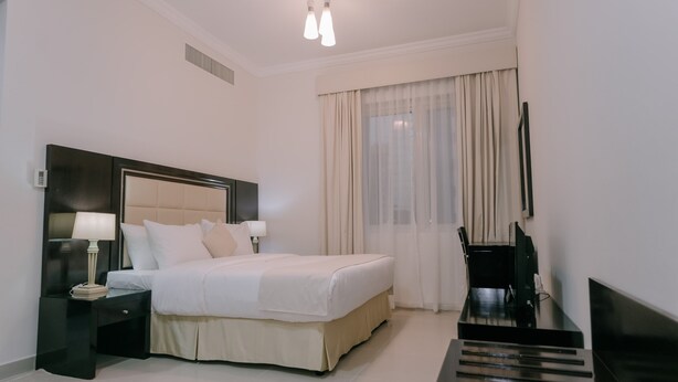 Una habitación de hotel moderna con una cama grande, mesitas de noche con lámparas, un televisor de pantalla plana y un ventanal con cortinas translúcidas.