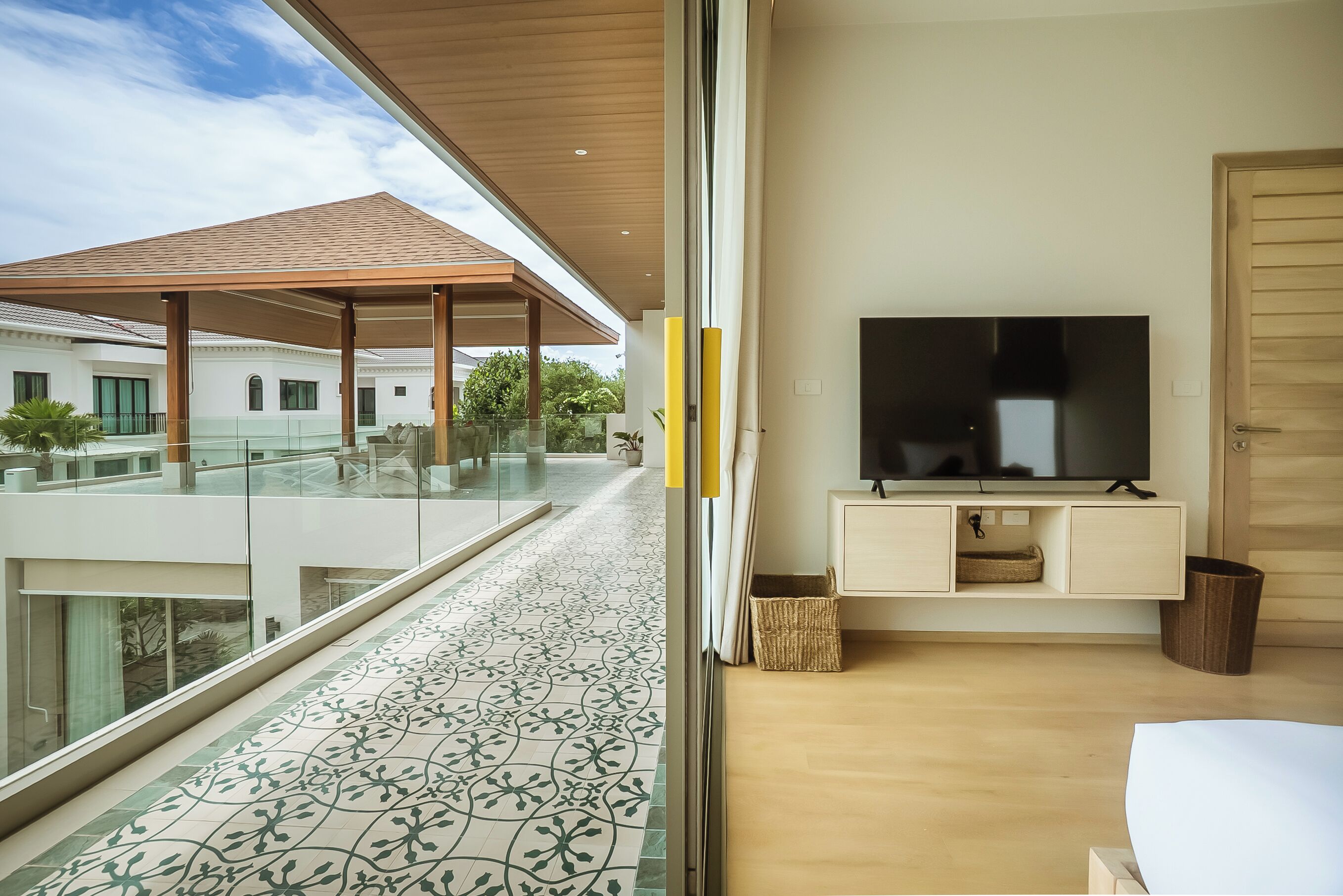 A modern living room with a large TV, wooden flooring, and a balcony with patterned tiles.
