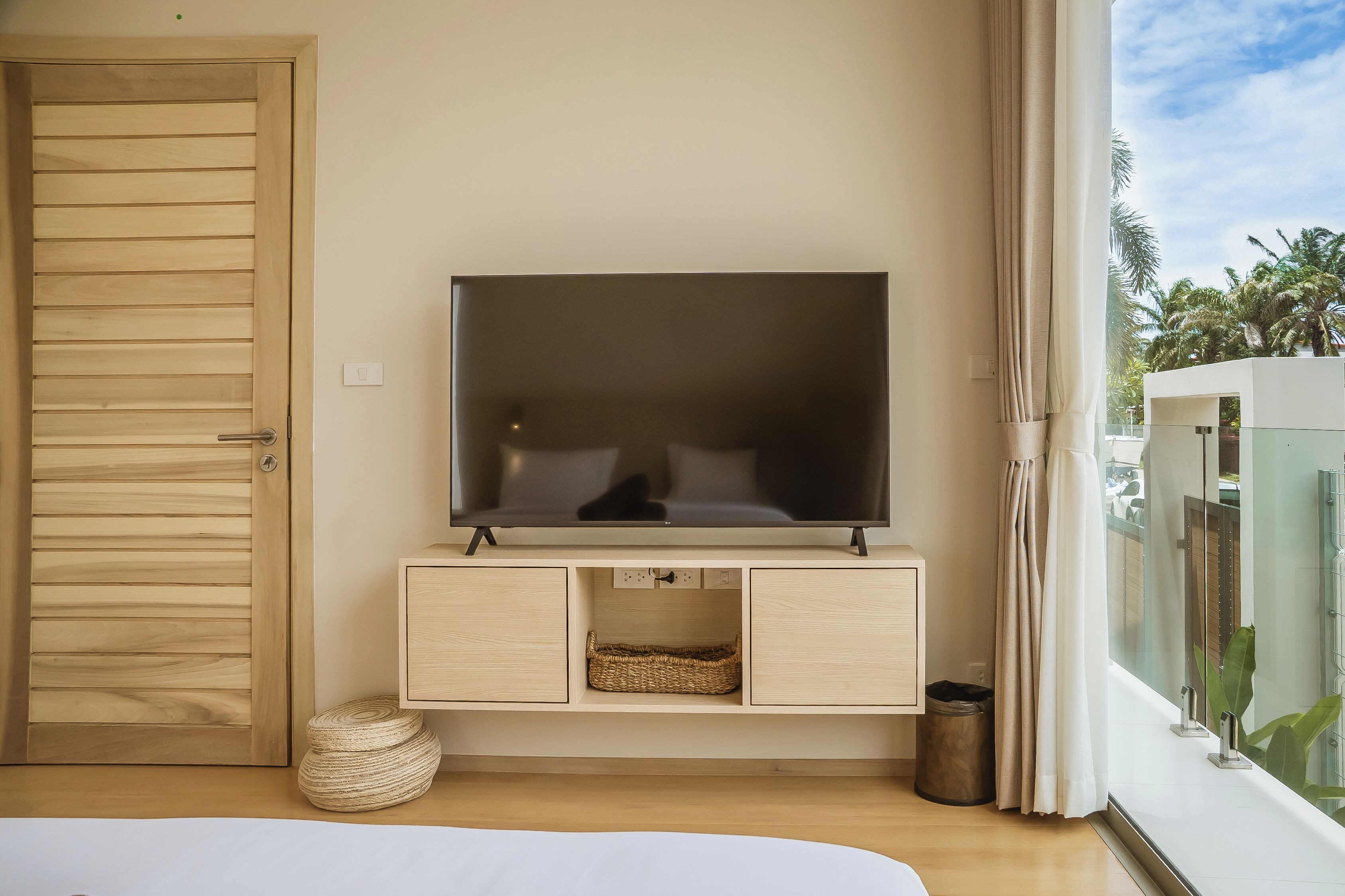 A modern room with a television on a wooden cabinet, a wicker basket, and a view of palm trees through large windows.