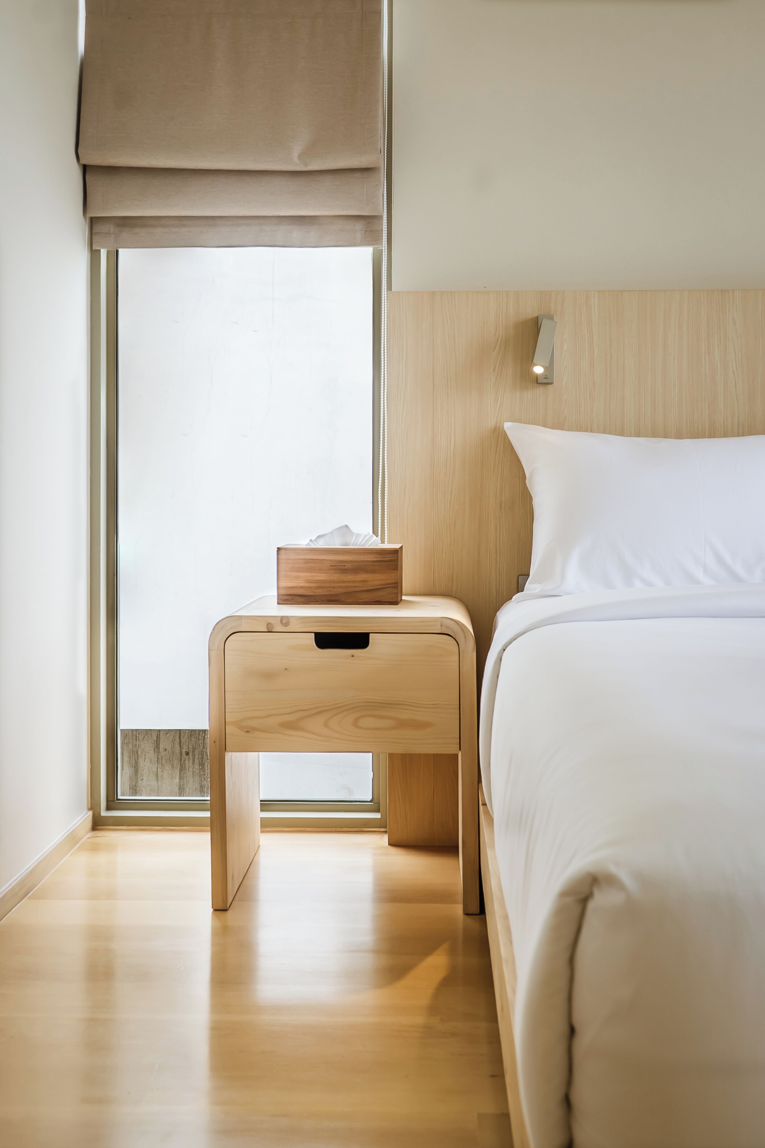 A minimalist bedroom with a wooden nightstand, a bedside table, a bed with white bedding, and a large window with a beige blind.