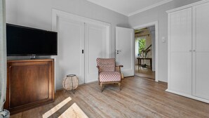 Una habitación amplia con pisos de madera, un televisor sobre un mueble de madera, una silla estampada y puertas blancas que dan a otras áreas.